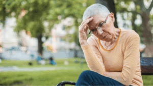 A lady with cropped gray hair and glasses puts her hand to her forehead, looking upset